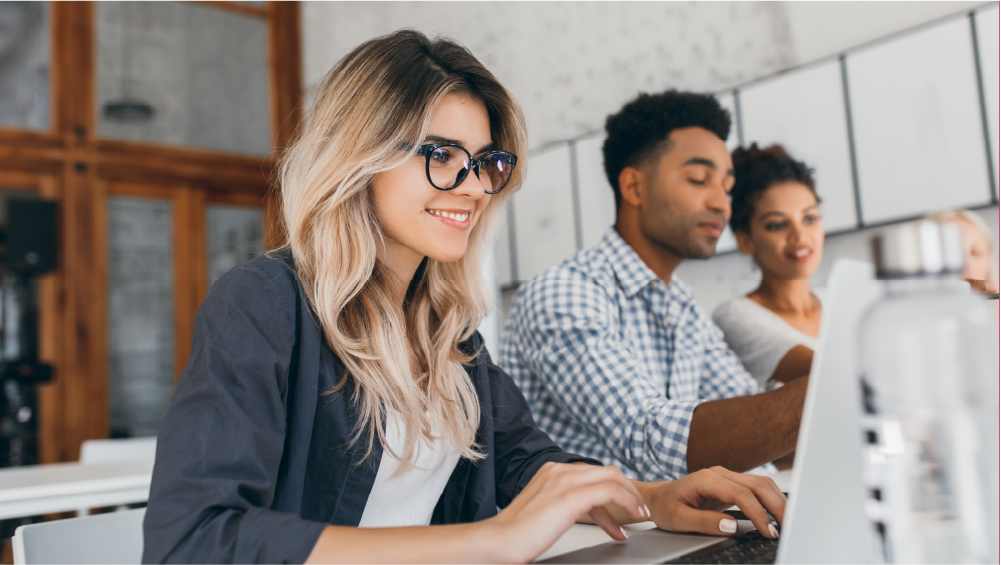 Foco em uma mulher loira usando um notebook e outras duas pessoas mais ao fundo também usando seus computadores.