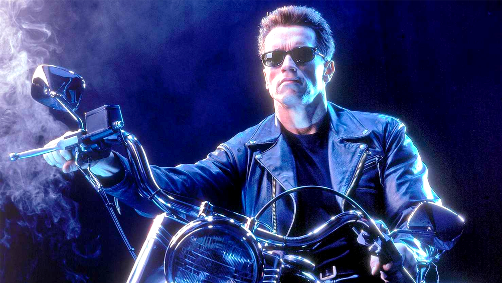 Numa cena noturna, o ator Arnold Schwarzenegger sentado em uma moto, usando óculos escuros e jaqueta de couro preto.