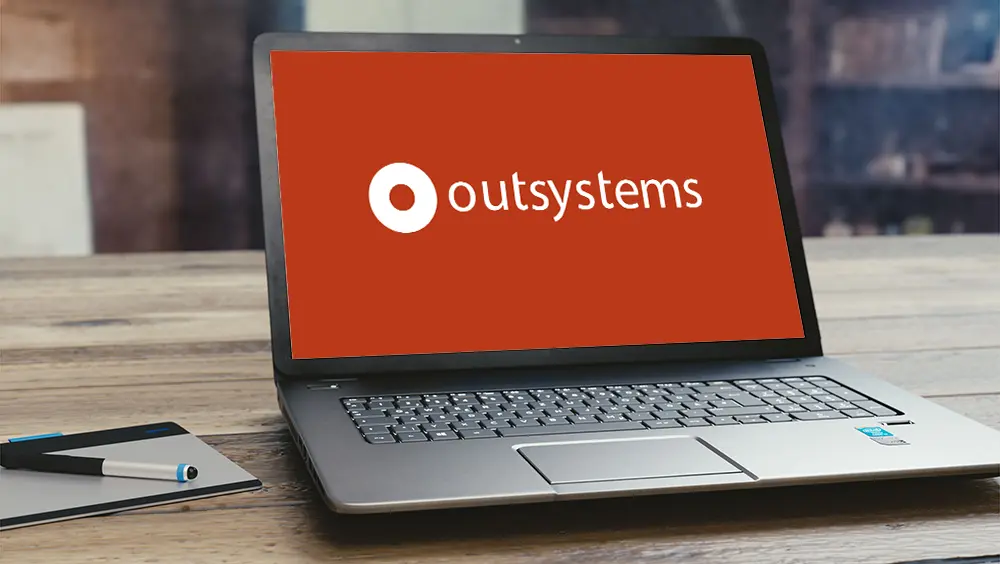 Um notebook aberto sob uma mesa de madeira, a tela está toda em vermelha com a logo do Outsystems ao centro