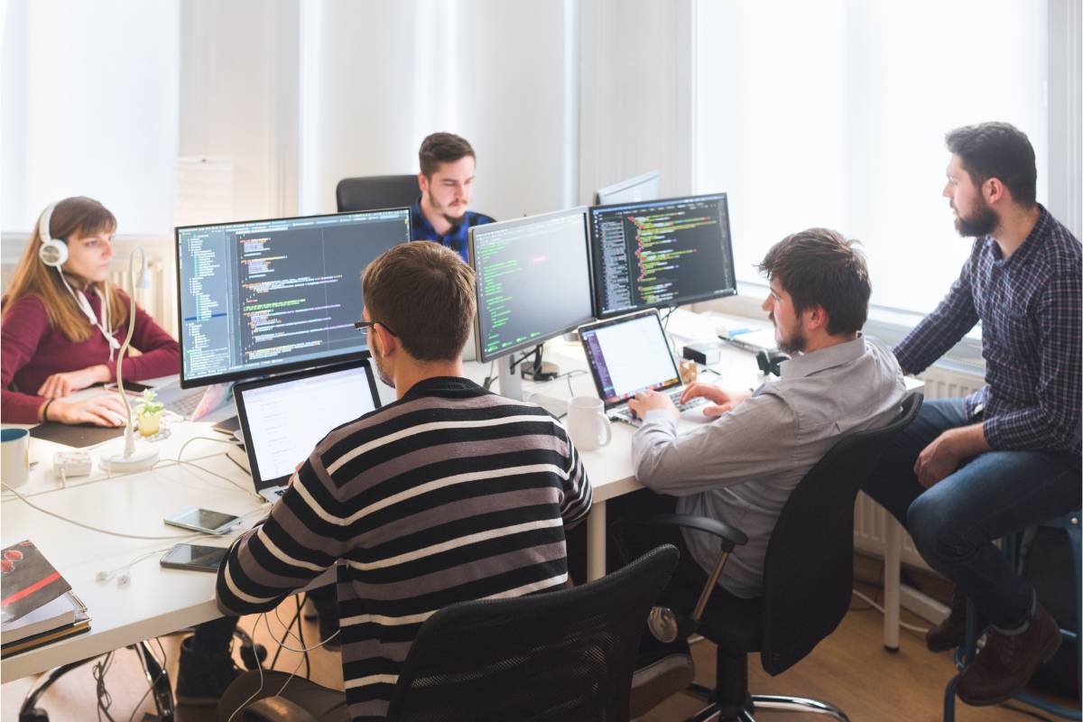 Team of developers working in a software factory, collaborating on development projects in an office environment with multiple monitors displaying code and programming tools.