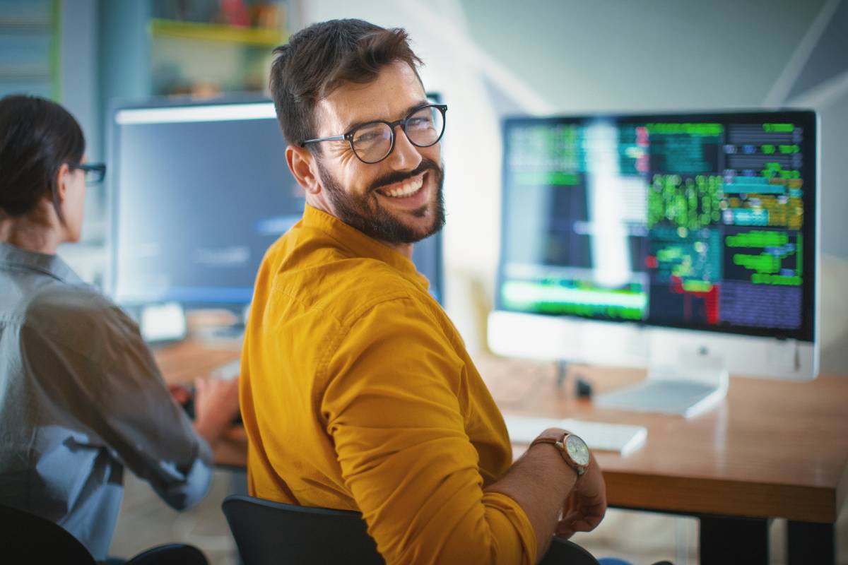 Profissional de TI sorridente sentado em frente a um monitor com códigos e gráficos, em um ambiente de escritório moderno.