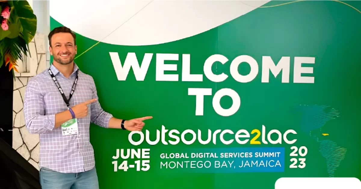 Juliano Haus, co-fundador da NextAge vestindo camisa xadrez e crachá, aponta para um grande painel verde com a mensagem "Welcome to Outsource2LAC – Global Digital Services Summit, Montego Bay, Jamaica, 2023". O evento é apoiado por organizações como IDB e ConnectAmericas.