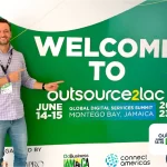 Juliano Haus, co-fundador da NextAge vestindo camisa xadrez e crachá, aponta para um grande painel verde com a mensagem "Welcome to Outsource2LAC – Global Digital Services Summit, Montego Bay, Jamaica, 2023". O evento é apoiado por organizações como IDB e ConnectAmericas.