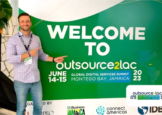 Juliano Haus, co-fundador da NextAge vestindo camisa xadrez e crachá, aponta para um grande painel verde com a mensagem "Welcome to Outsource2LAC – Global Digital Services Summit, Montego Bay, Jamaica, 2023". O evento é apoiado por organizações como IDB e ConnectAmericas.