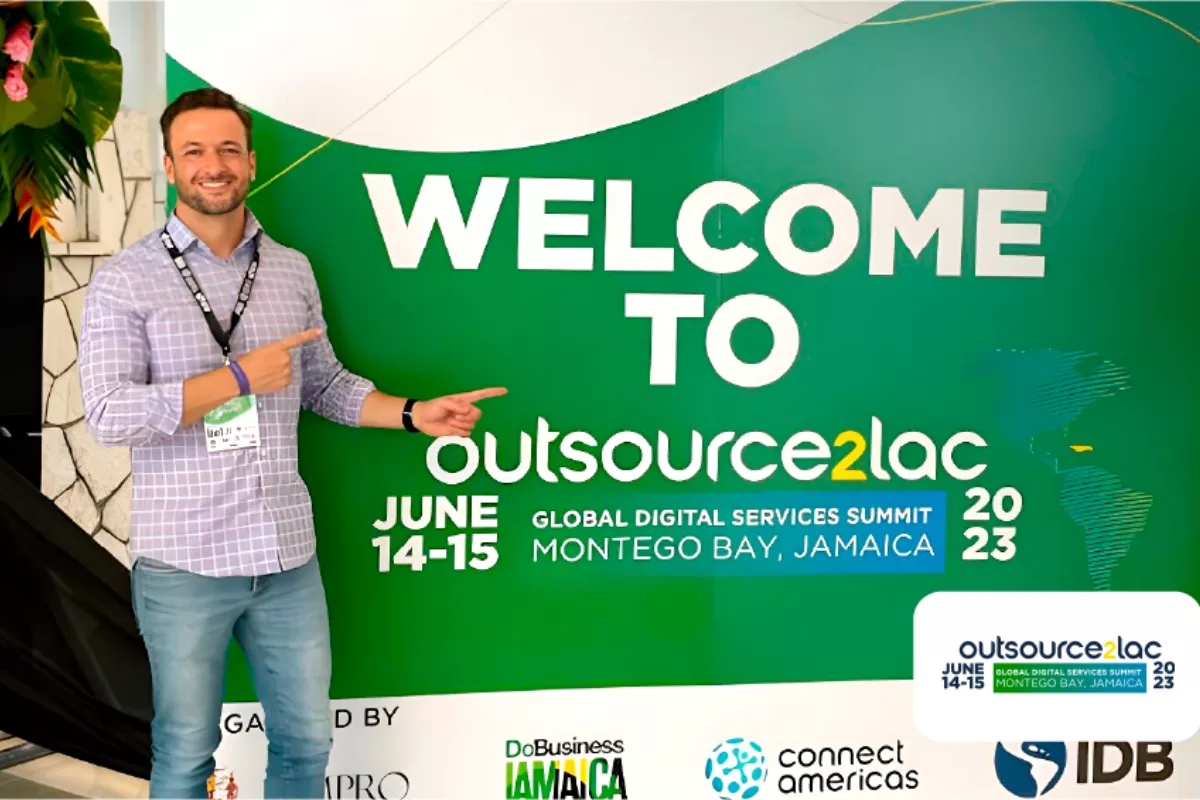 Juliano Haus, co-fundador da NextAge vestindo camisa xadrez e crachá, aponta para um grande painel verde com a mensagem "Welcome to Outsource2LAC – Global Digital Services Summit, Montego Bay, Jamaica, 2023". O evento é apoiado por organizações como IDB e ConnectAmericas.