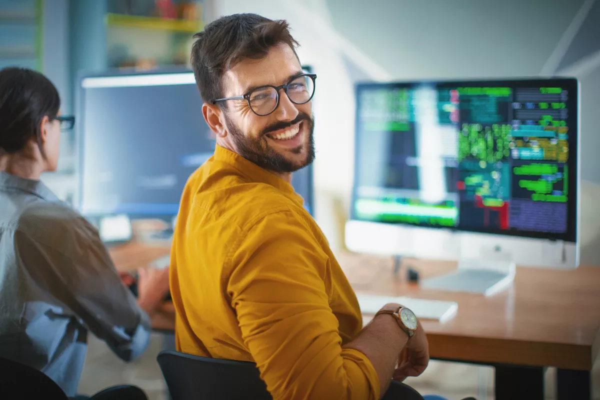 Smiling man wearing glasses and a yellow shirt in a tech environment, sitting in front of a computer screen displaying code. In the background, a colleague is working at another monitor. The image conveys a productive and collaborative atmosphere, representing the application of Lean Methodology for efficiency and high performance in IT teams.