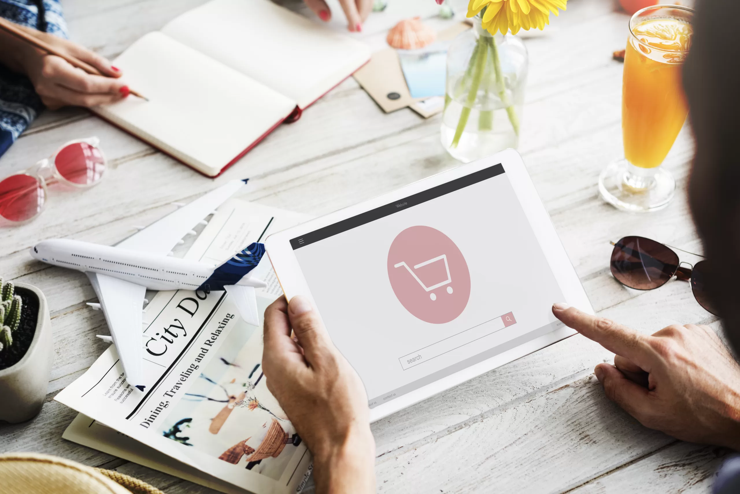 Person using tablet with e-commerce icon; table with juice, sunglasses, flowers, and model airplane.