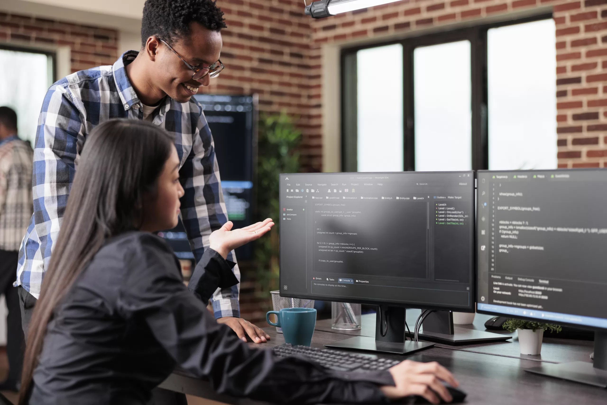 Programador em pé explicando código para uma colega sentada, enquanto analisam uma interface de desenvolvimento exibida em dois monitores.