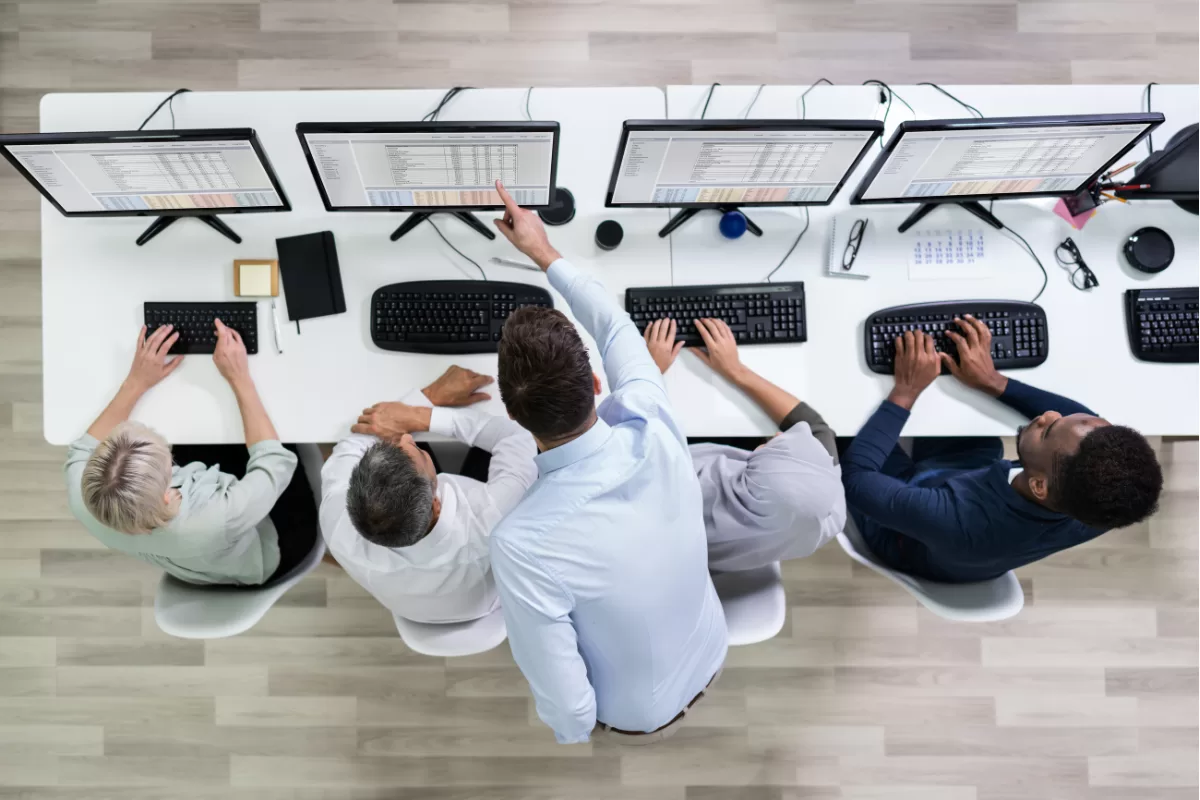 Equipe de profissionais de TI trabalhando lado a lado em frente a quatro monitores, analisando dados e colaborando em um projeto para otimizar operações. 