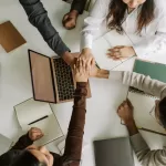 Grupo de colegas unindo as mãos sobre uma mesa com laptops e cadernos, simbolizando trabalho em equipe e colaboração.
