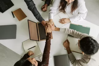 Grupo de colegas unindo as mãos sobre uma mesa com laptops e cadernos, simbolizando trabalho em equipe e colaboração.
