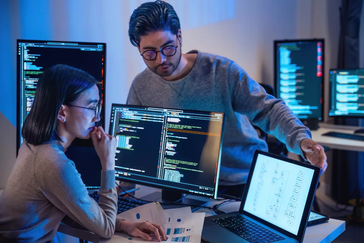 Dois desenvolvedores de software trabalhando juntos em frente a vários monitores com código, um mostrando um diagrama em um tablet, representando colaboração em programação e design de sistemas.