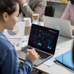 Profissional analisando gráficos e relatórios em um notebook durante reunião de equipe, representando validão de entregas