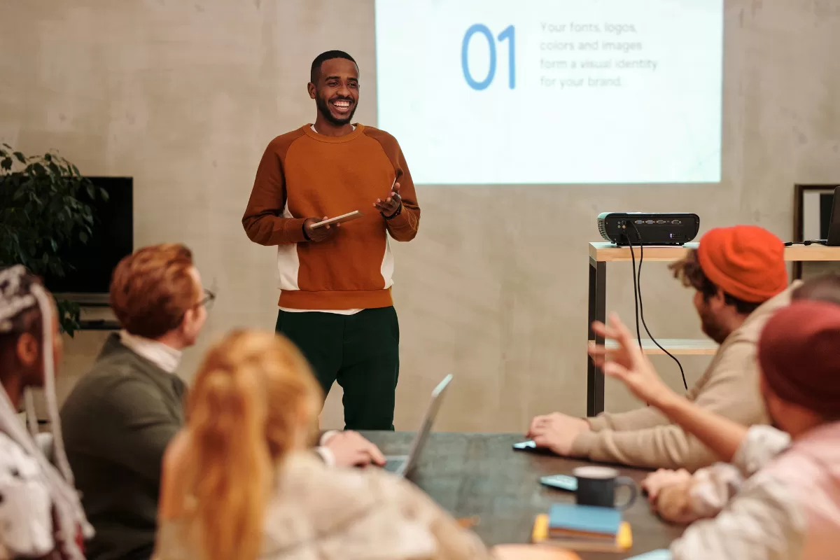 Smiling man standing and giving a presentation to a group of people in a room, with a projector displaying a slide behind him, representing tips for presenting your IT project