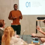 Homem em pé sorridente fazendo uma apresentação para um grupo de pessoas em uma sala, com um projetor exibindo um slide atrás dele, representando apresentação de métricas