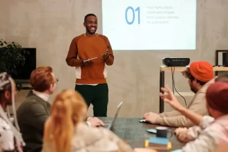Homem em pé sorridente fazendo uma apresentação para um grupo de pessoas em uma sala, com um projetor exibindo um slide atrás dele, representando apresentação de métricas