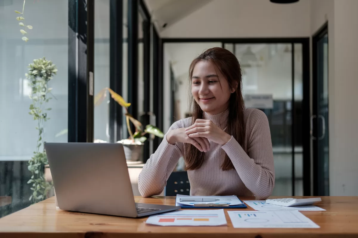 Mulher sentada à mesa em frente a um laptop, sorrindo enquanto participa de uma reunião online, com documentos e calculadora sobre a mesa.