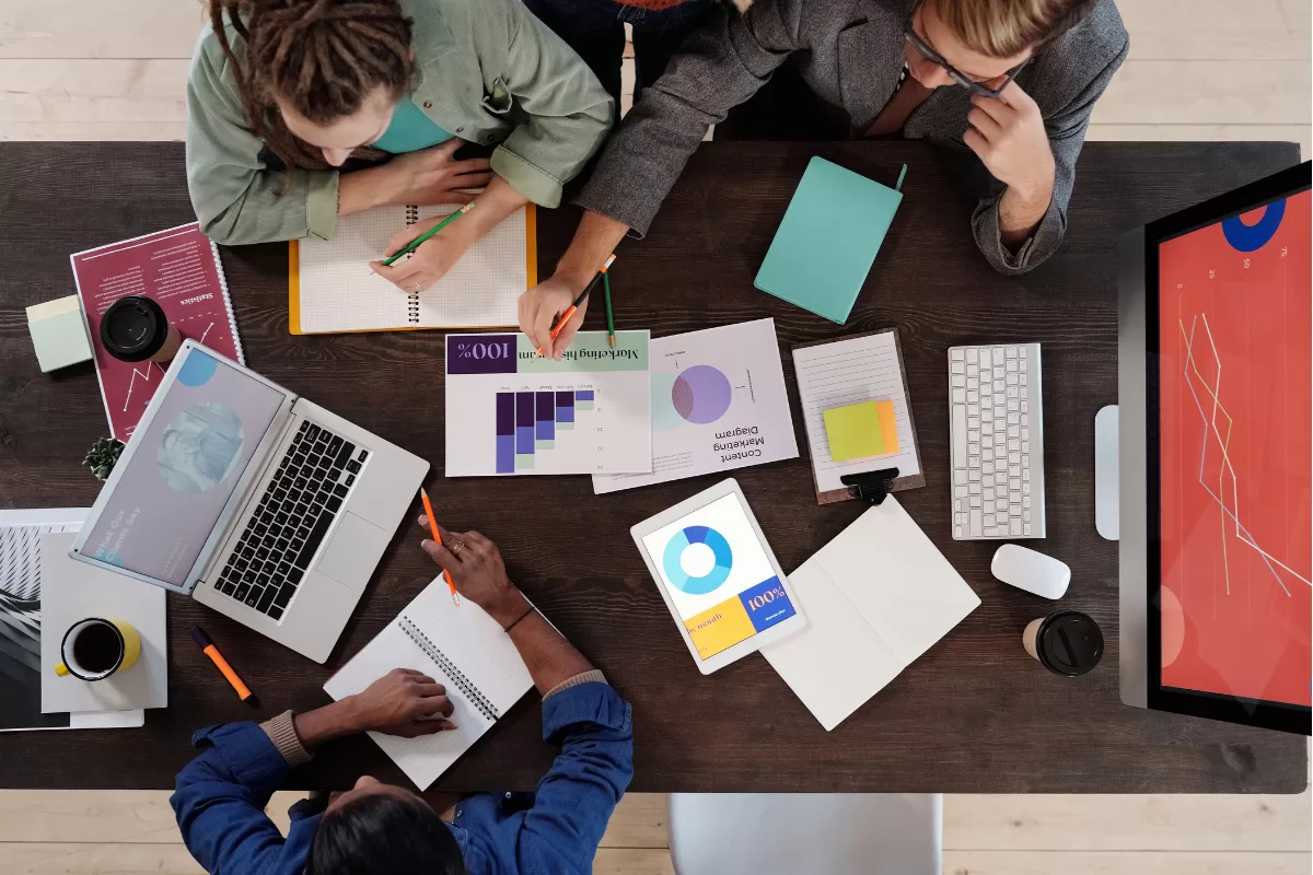 Top-down view of a team reviewing charts and project plans at a shared desk with a laptop, tablet, and reports.