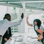 Equipe de TI celebrando um bom resultado no escritório, com colegas dando high-five ao redor de uma mesa com notebooks, representando centralizar ou terceirizar equipes de TI