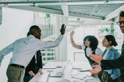 Equipe de TI celebrando um bom resultado no escritório, com colegas dando high-five ao redor de uma mesa com notebooks, representando centralizar ou terceirizar equipes de TI