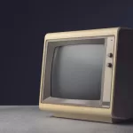 Monitor CRT vintage bege dos anos 80/90 com tela apagada sobre mesa de madeira com fundo escuro, representando tecnologia desatualizada