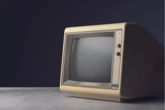 Monitor CRT vintage bege dos anos 80/90 com tela apagada sobre mesa de madeira com fundo escuro, representando tecnologia desatualizada