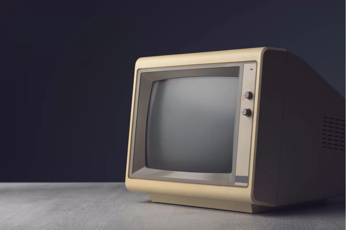 Monitor CRT vintage bege dos anos 80/90 com tela apagada sobre mesa de madeira com fundo escuro, representando tecnologia desatualizada