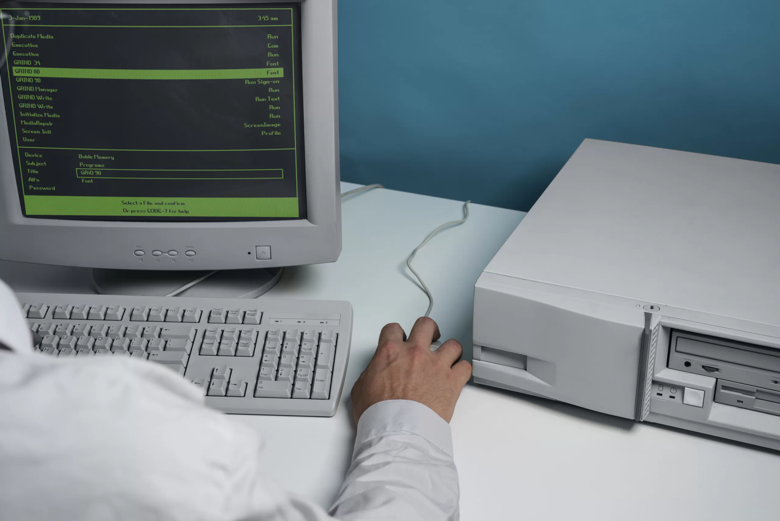 Computador desktop antigo com monitor CRT exibindo interface verde de sistema legado, teclado branco e CPU, com mão de pessoa interagindo com o equipamento