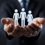 Mãos de uma pessoa em um terno segurando cuidadosamente três pequenas figuras brancas estilizadas que representam pessoas, sugerindo integração ou cuidado com a equipe.