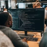 Monitor exibindo código de programação em primeiro plano com equipe de desenvolvimento trabalhando colaborativamente ao fundo em ambiente de escritório, representando modelos de contratação de TI