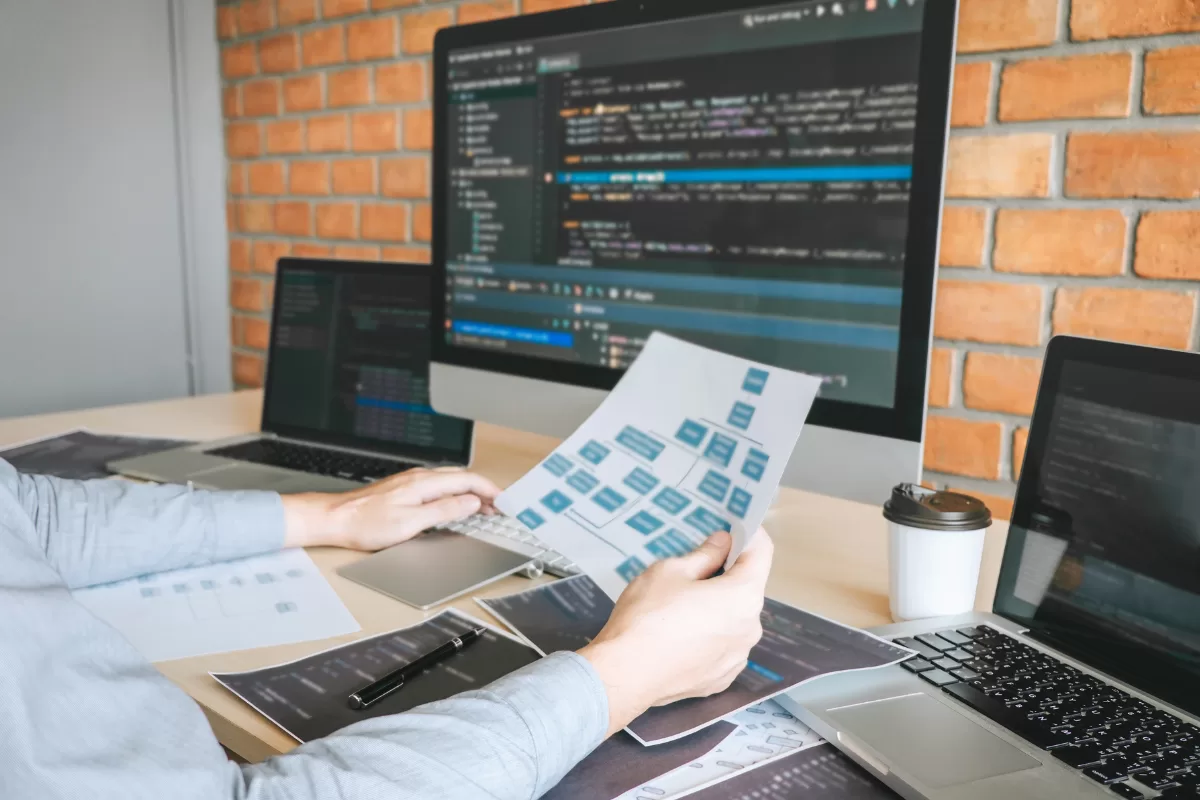 Person holding document with blue flowchart diagram in front of monitor displaying code, on desk with laptop, tablet and coffee
