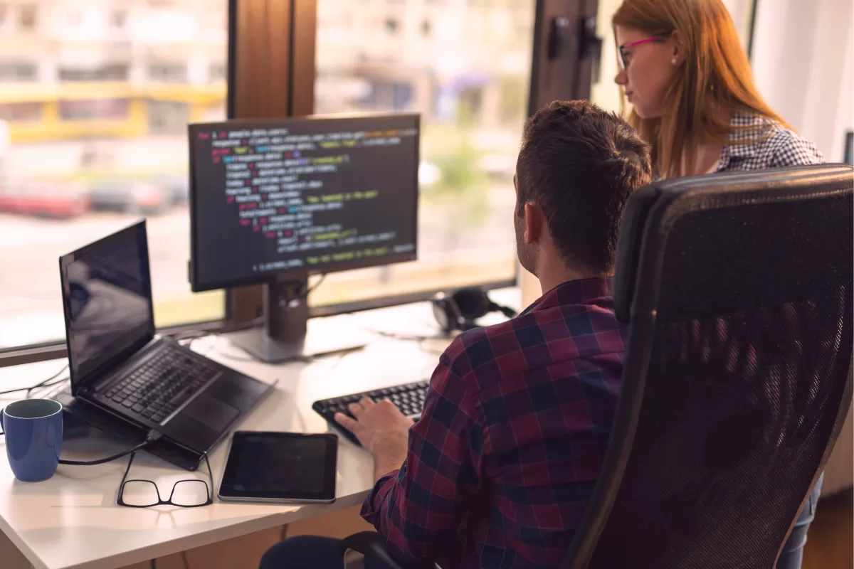 Two developers working together in front of monitor displaying code, man sitting typing and woman standing observing