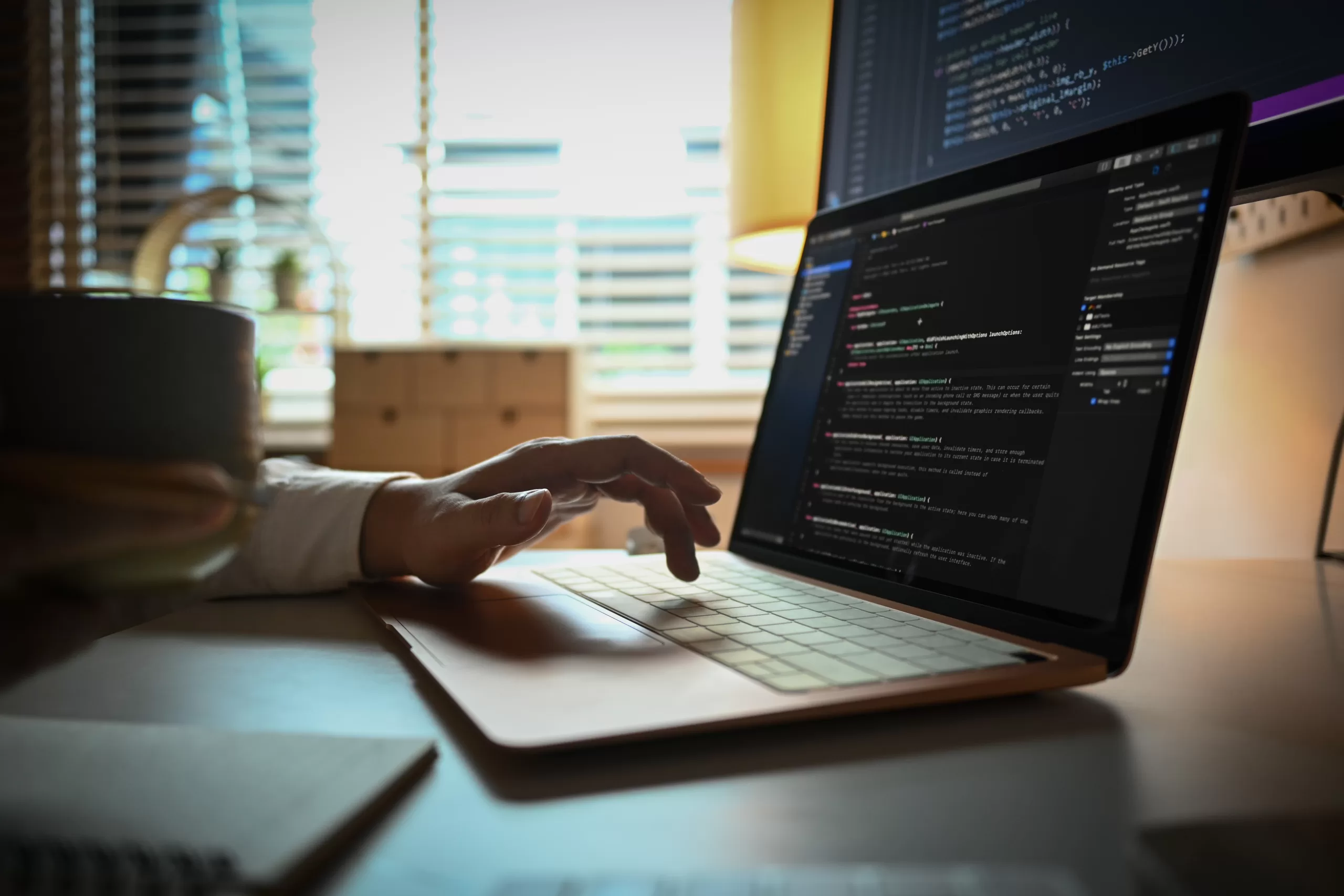 Close-up of a programmer's hand typing on a laptop with code displayed on an external screen, representing refactoring and code maintenance.