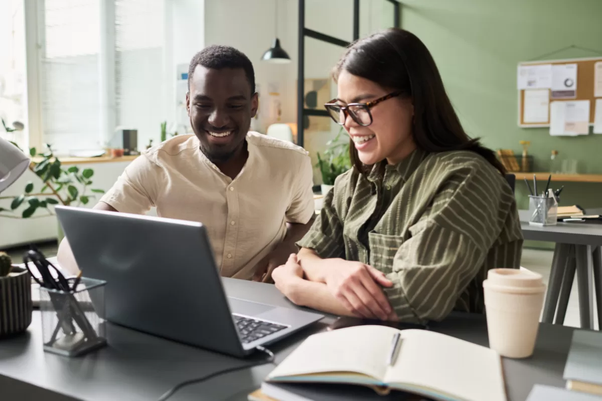 Duas pessoas, um homem negro e uma mulher asiática, sorrindo enquanto olham juntas para a tela de um laptop em um escritório moderno.