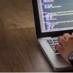 Hand typing on a laptop with a code editor open on a wooden desk.