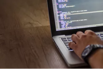 Hand typing on a laptop with a code editor open on a wooden desk.
