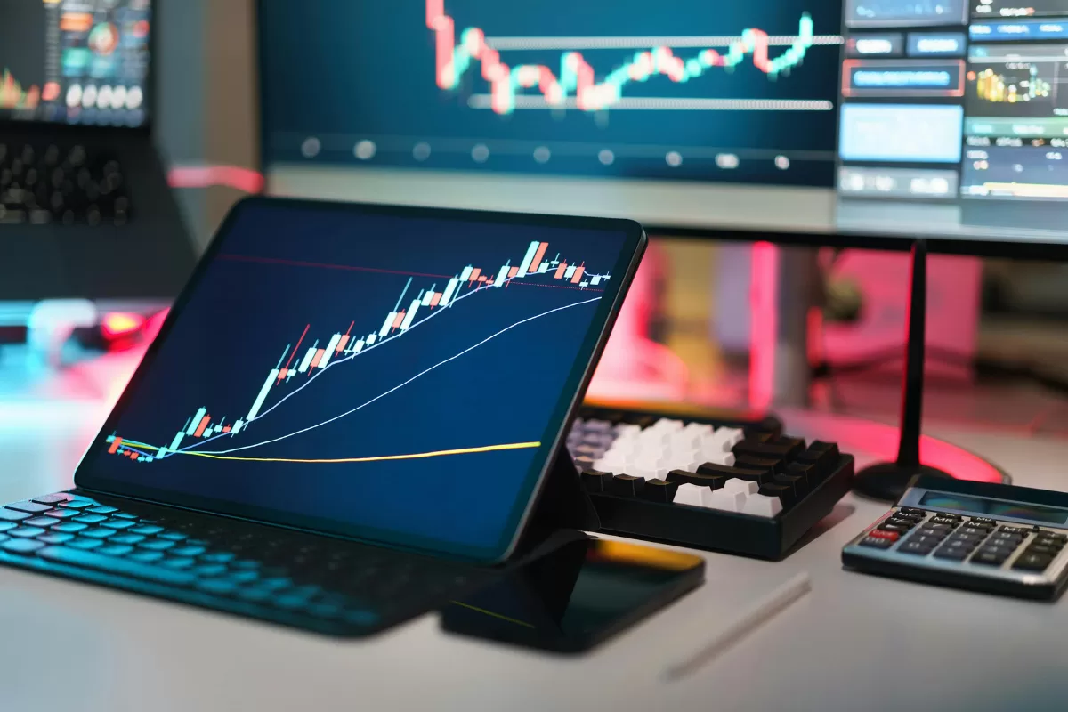 Tablet exibindo gráficos de candlestick financeiros em ambiente de trading com monitor ao fundo mostrando dados do mercado, teclado e calculadora sobre mesa de trabalho, representando Core Banking