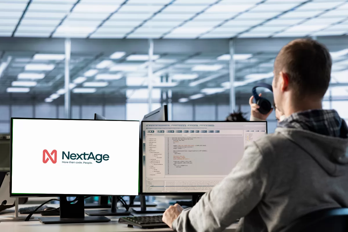 Developer in modern office working with dual monitors, one displaying NextAge logo with tagline 'More than code. People.' and another showing programming code