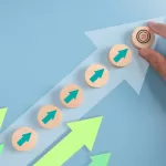 Uma mão coloca um bloco de madeira com um alvo em cima de uma escada de blocos de madeira com setas verdes, simbolizando o progresso da carreira em direção a um objetivo. O fundo é azul claro com setas de crescimento. Isso representa gestão de carreira para TI
