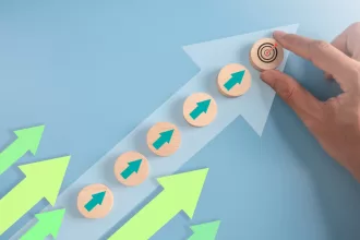 Uma mão coloca um bloco de madeira com um alvo em cima de uma escada de blocos de madeira com setas verdes, simbolizando o progresso da carreira em direção a um objetivo. O fundo é azul claro com setas de crescimento. Isso representa gestão de carreira para TI