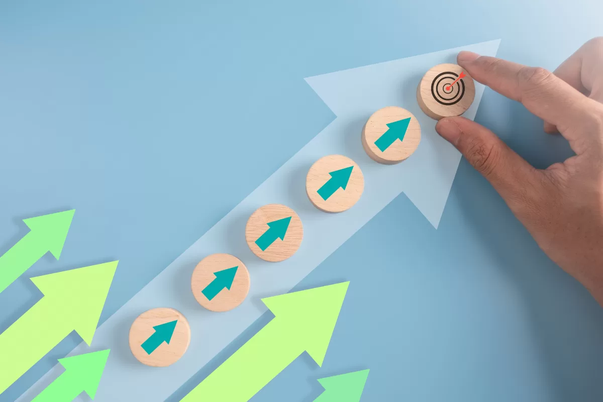 Uma mão coloca um bloco de madeira com um alvo em cima de uma escada de blocos de madeira com setas verdes, simbolizando o progresso da carreira em direção a um objetivo. O fundo é azul claro com setas de crescimento. Isso representa gestão de carreira para TI