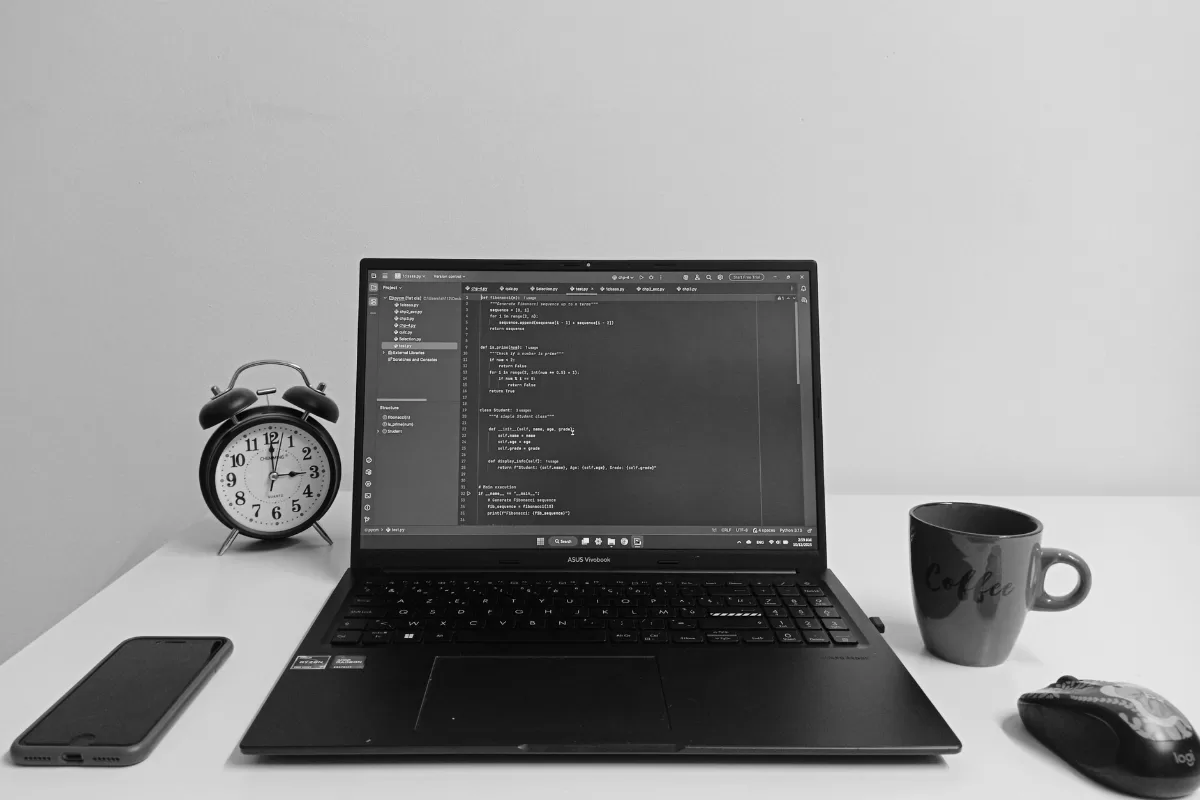 Uma foto em preto e branco de um laptop aberto exibindo código em um editor de texto, ao lado de um despertador, um smartphone, um mouse e uma caneca de café. O ambiente sugere trabalho noturno ou foco em programação.