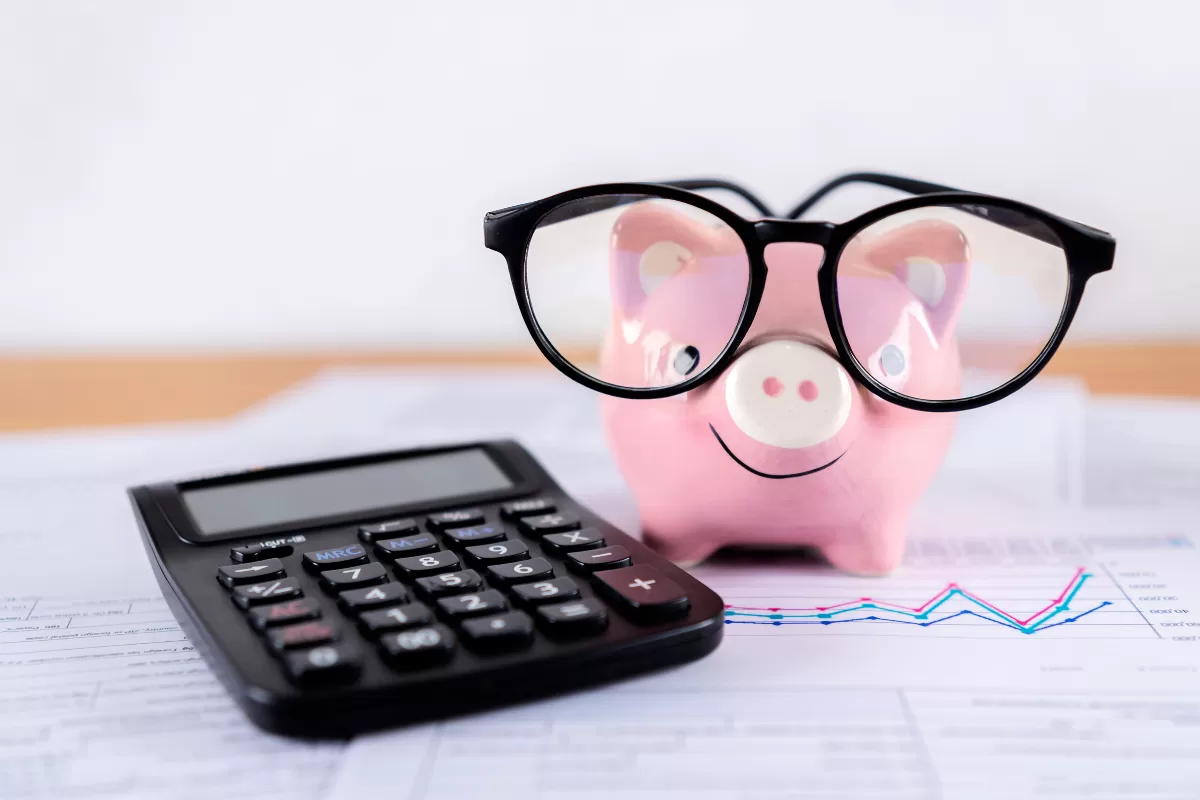 Cofre de porquinho rosa usando óculos ao lado de calculadora e gráficos financeiros sobre mesa, representando planejamento de orçamento