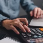 Mãos de profissional usando calculadora enquanto trabalha no laptop, simbolizando cálculo de custos e planejamento financeiro de TI