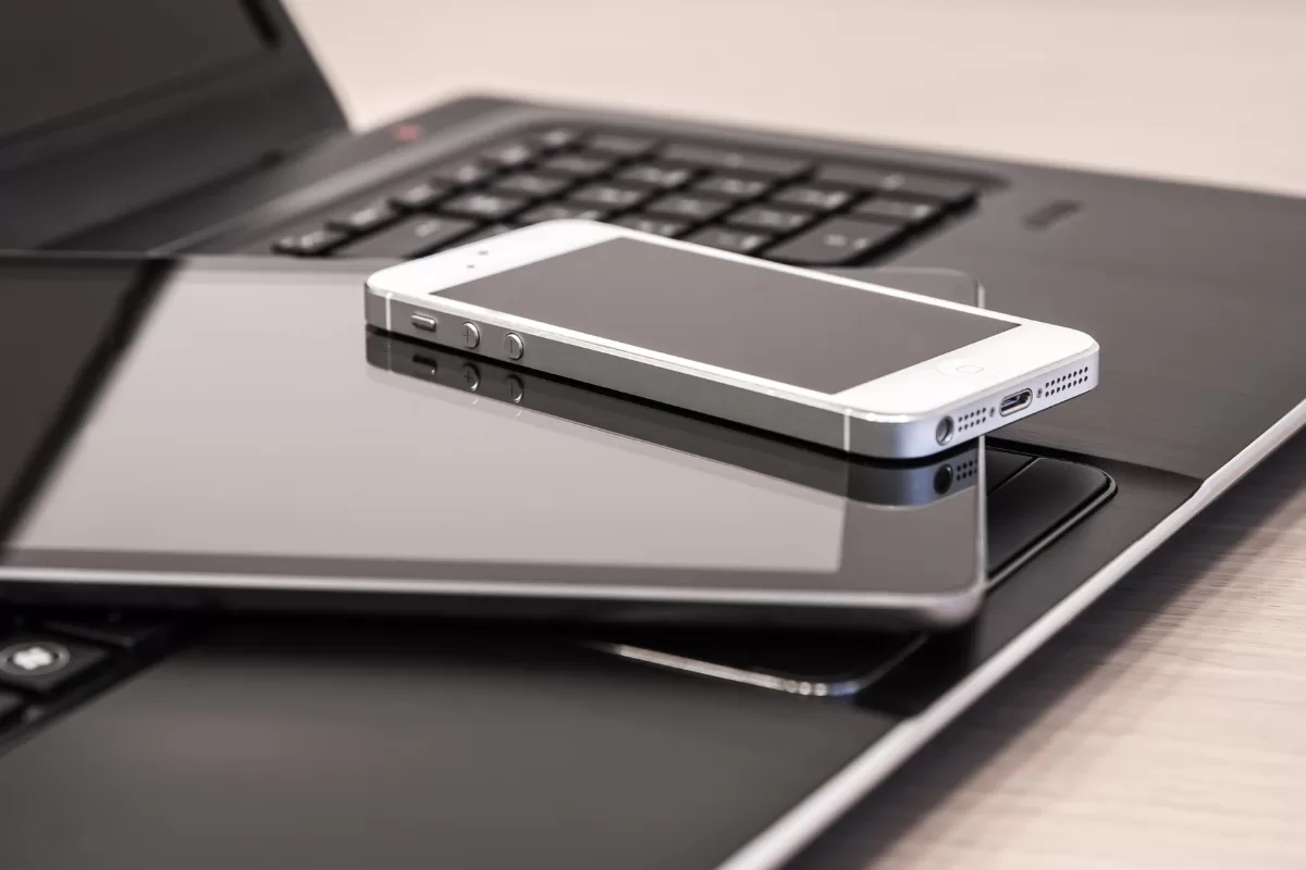 An overhead, close-up view of a collection of modern personal electronic devices—a silver smartphone, a dark tablet, and a black laptop—stacked or placed together on a wooden surface.