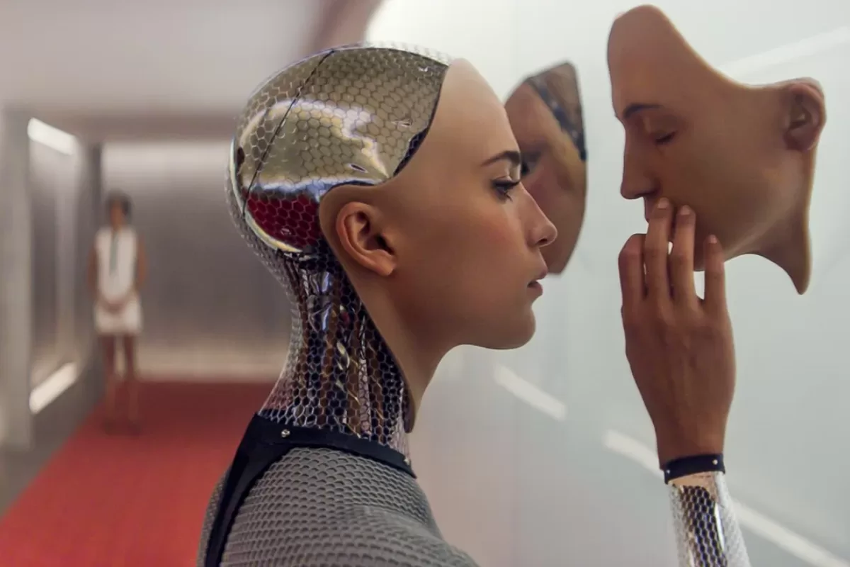 Androide feminina observa de perto um rosto artificial humano em uma parede, cena do filme Ex Machina.