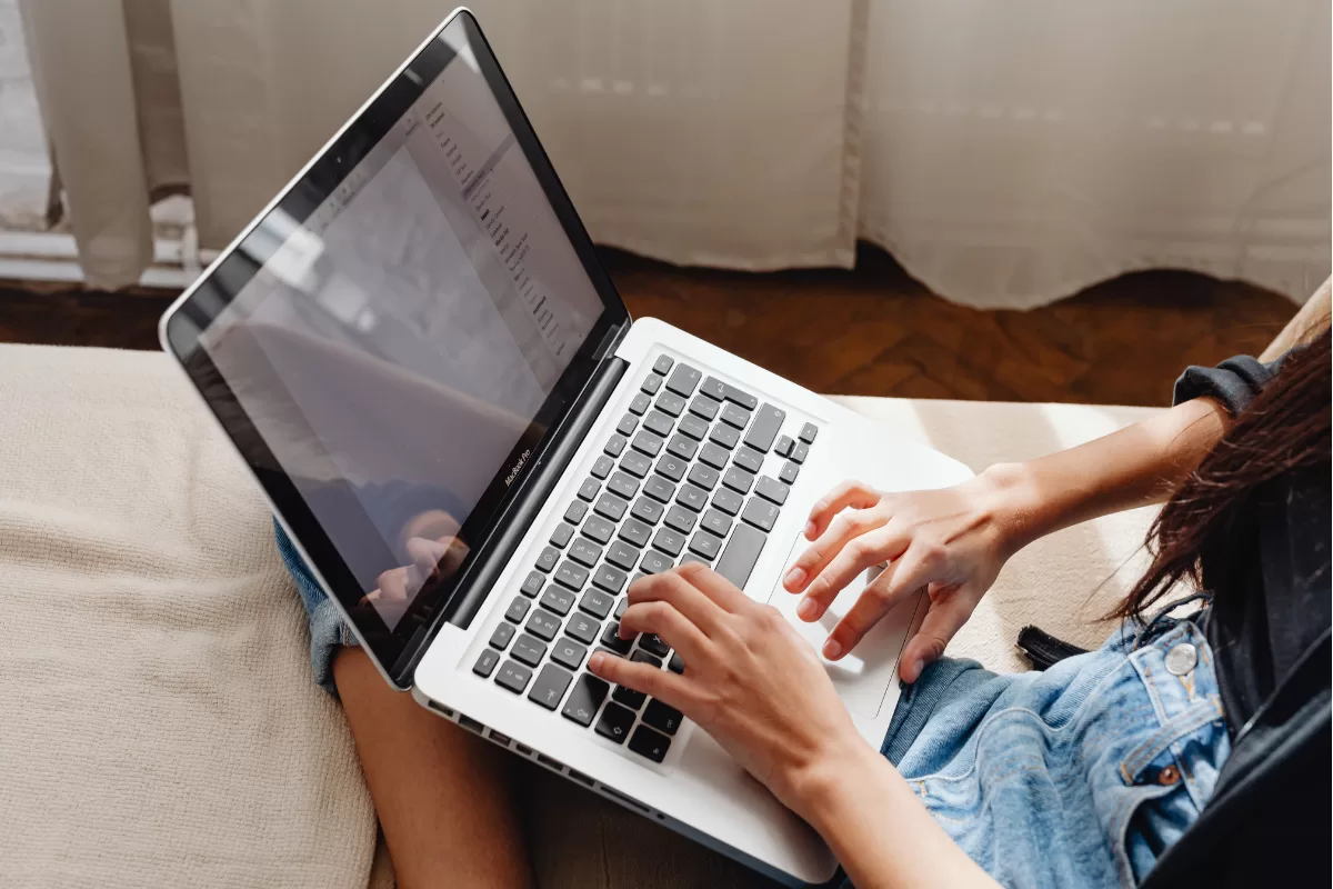 Person typing on silver laptop on beige couch, with screen displaying programming code