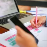 Mão segurando um smartphone e outra anotando em rascunhos de interface; ao fundo, notebook com wireframes de telas mobile na tela.