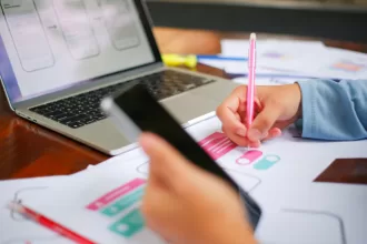 Mão segurando um smartphone e outra anotando em rascunhos de interface; ao fundo, notebook com wireframes de telas mobile na tela.