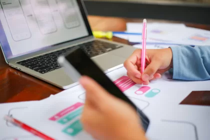 Mão segurando um smartphone e outra anotando em rascunhos de interface; ao fundo, notebook com wireframes de telas mobile na tela.